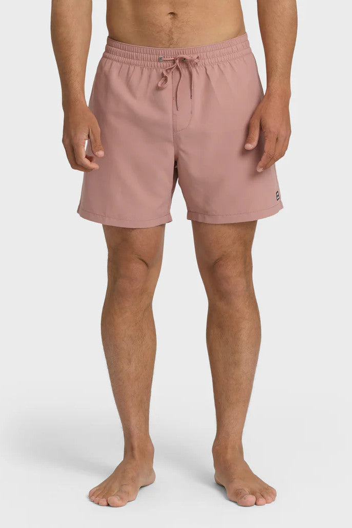 A man standing and posing in a pair of rose pink boardshorts with a side pocket, featuring an elastic waist closure.