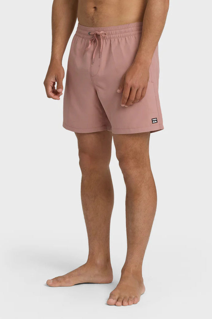 A man standing and posing in a pair of rose pink boardshorts with a side pocket, featuring an elastic waist closure.