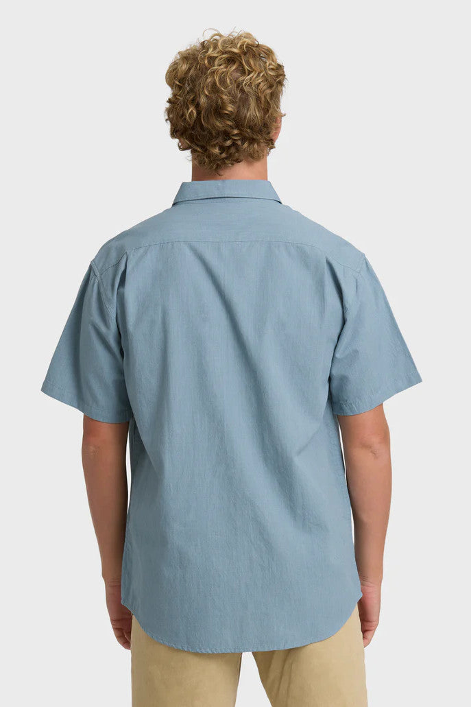 Back view of a man wearing a solid, blue short-sleeve button-up collared shirt on a white background