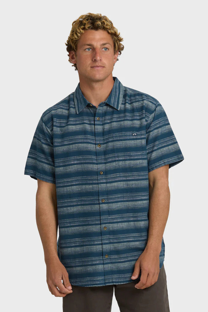 A man wearing a striped short-sleeved woven shirt with a collar and button-up closure, featuring a chest pocket. The shirt is primarily dark blue with faded cream stripes.