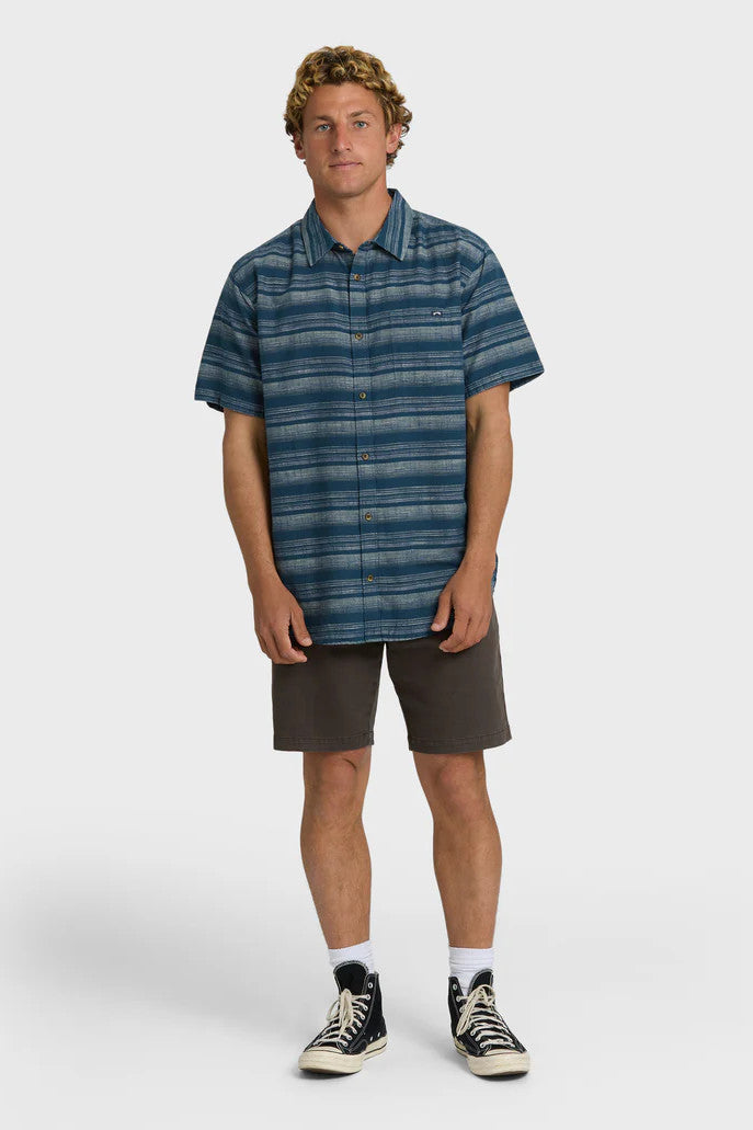 A man wearing a striped short-sleeved woven shirt with a collar and button-up closure, featuring a chest pocket. The shirt is primarily dark blue with faded cream stripes.