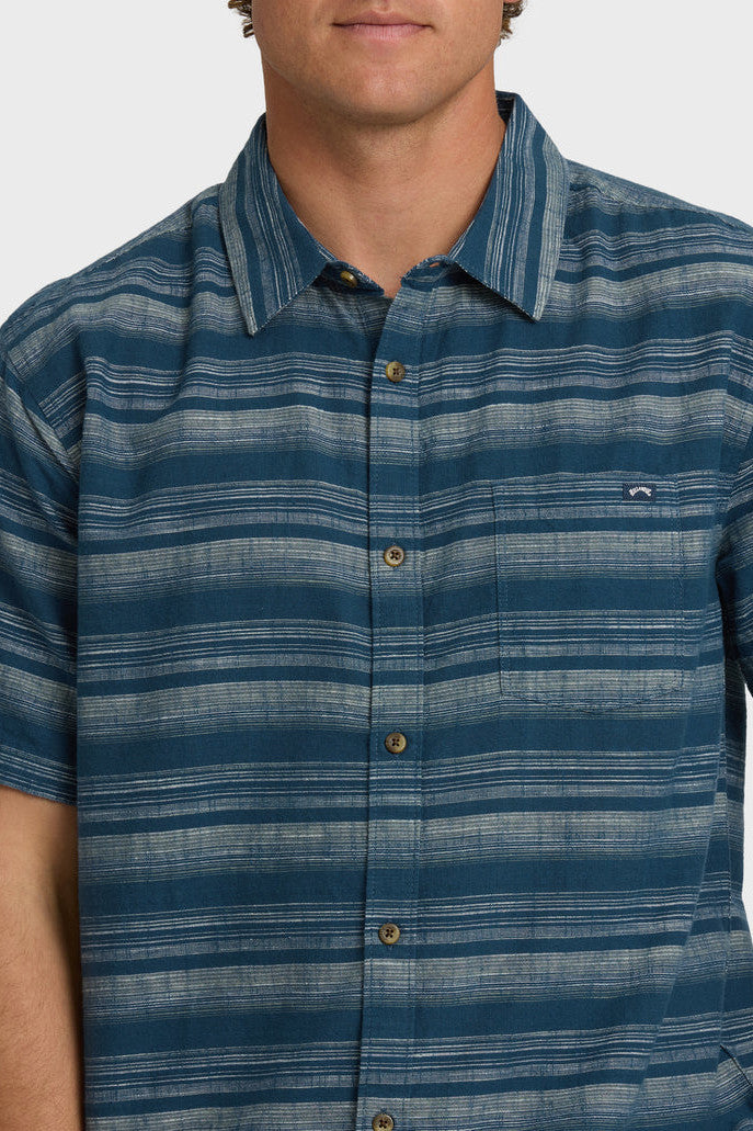 A man wearing a striped short-sleeved woven shirt with a collar and button-up closure, featuring a chest pocket. The shirt is primarily dark blue with faded cream stripes.