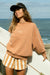 Woman holding a surfboard by the ocean wearing a brown sweatshirt and striped shorts.