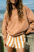 Woman holding a surfboard by the ocean wearing a brown sweatshirt and striped shorts.