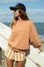 Woman holding a surfboard by the ocean wearing a brown sweatshirt and striped shorts.