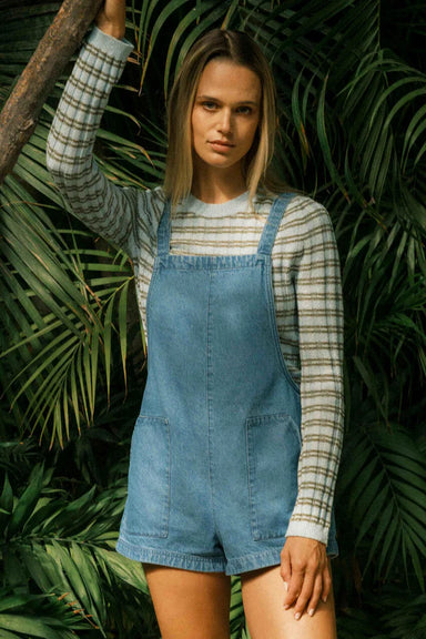 Woman wearing denim overalls and a striped sweater standing among green foliage