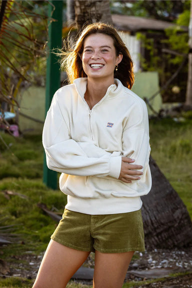 Woman wearing a cream-colored hoodie and green shorts standing outdoors with trees and grass in the background.