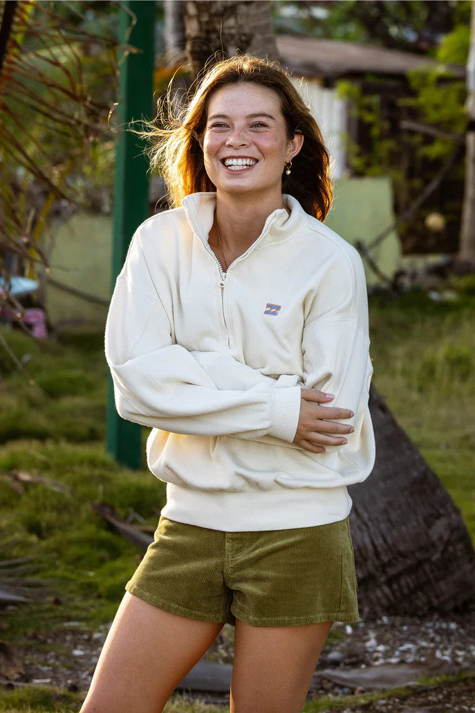 Woman wearing a cream-colored hoodie and green shorts standing outdoors with trees and grass in the background.