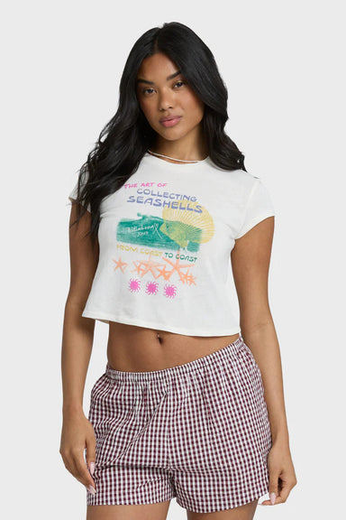 Woman wearing a white graphic t-shirt that says "the art of collecting seashells" and checkered shorts on a white background