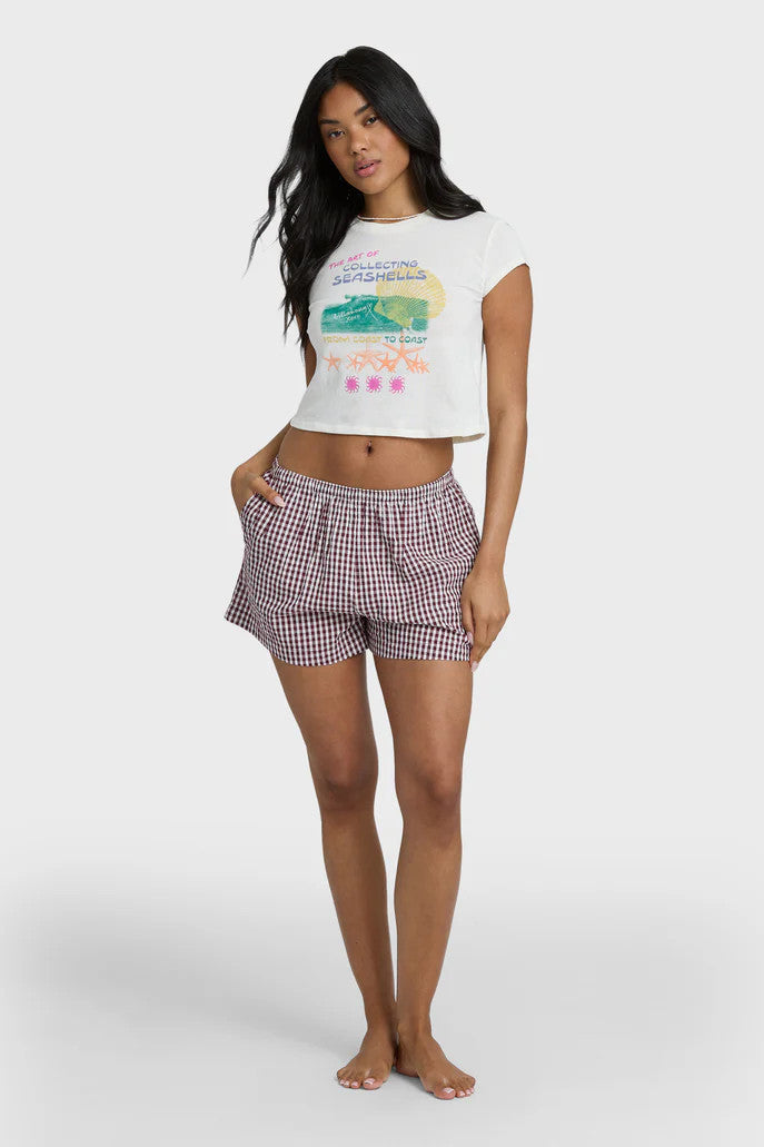 Woman wearing a white graphic t-shirt that says "the art of collecting seashells" and checkered shorts on a white background
