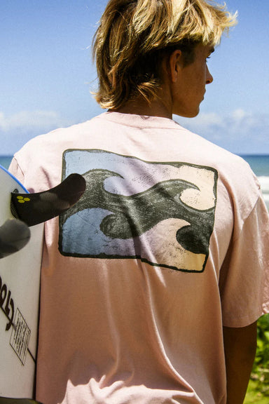 Man wearing a light pink t-shirt with a wave graphic design on the back, holding a surfboard in front of a blue sky