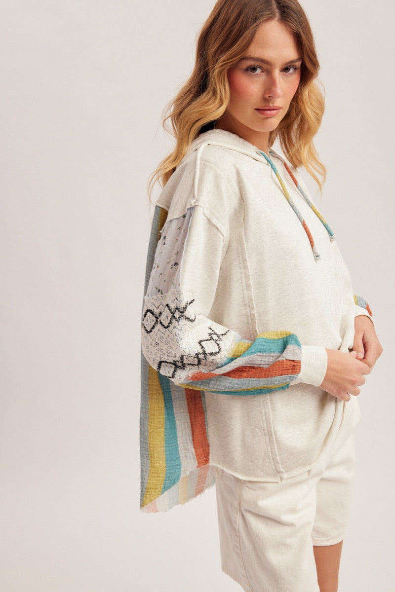 Woman wearing an oatmeal hoodie with colorful stripes and design, standing against a plain background