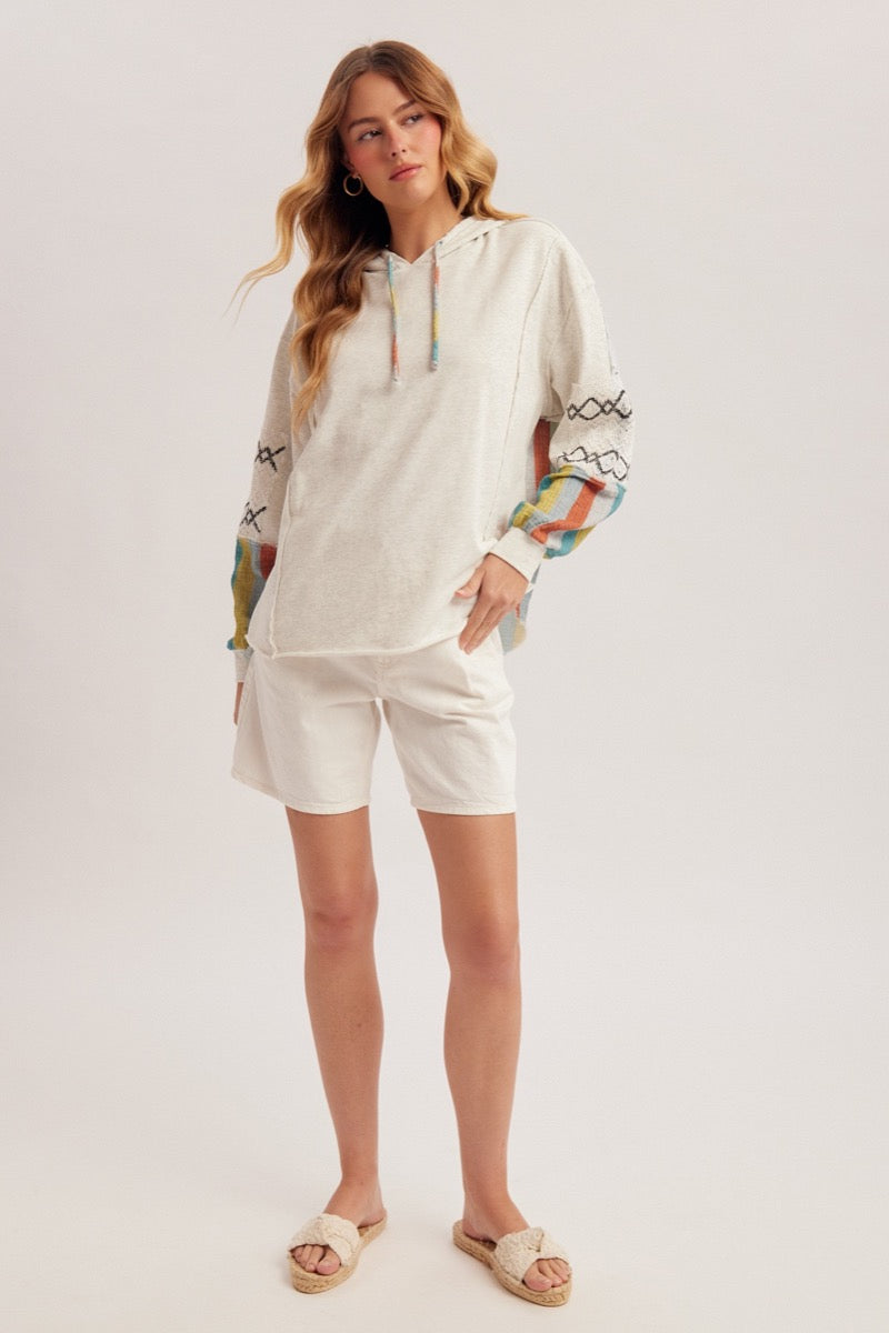 Woman wearing an oatmeal hoodie with colorful designs and white shorts on a light gray background