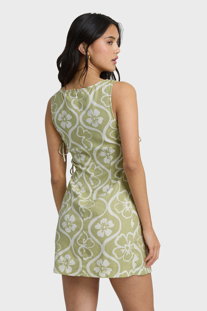 Back view of a woman wearing a green and white patterned dress with side ties on a white background