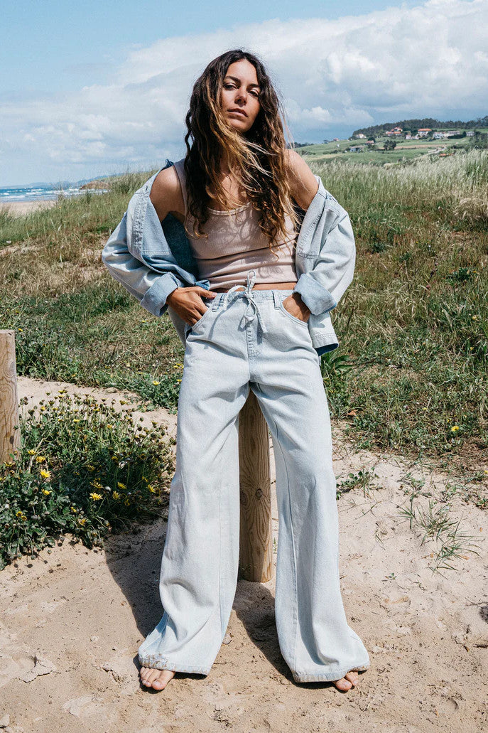 Woman standing by the dunse on a beach with ocean and sky in the background. Wearing a beige tank top, light denim shirts, and matching light denim pants