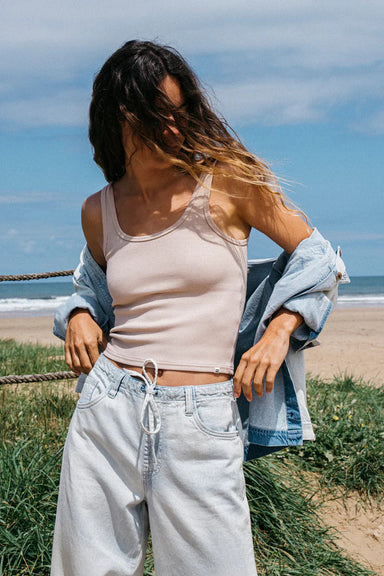 Woman standing on a beach wearing a beige tank top and light blue jeans with a denim shirt over her shoulder.