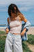 Woman standing on a beach wearing a beige tank top and light blue jeans with a denim shirt over her shoulder.