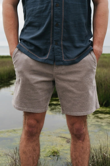 Person wearing a blue shirt and beige shorts standing in a natural setting with water and grass.