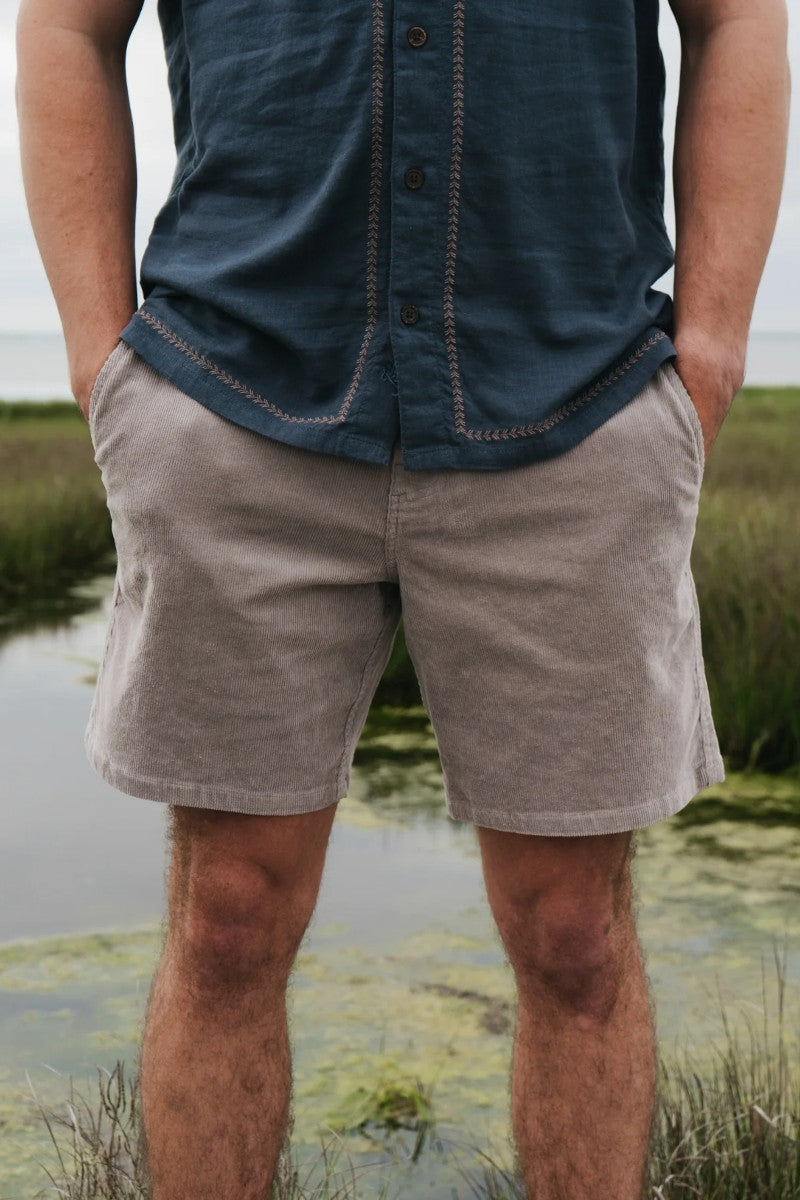 Person wearing a blue shirt and beige shorts standing in a natural setting with water and grass.