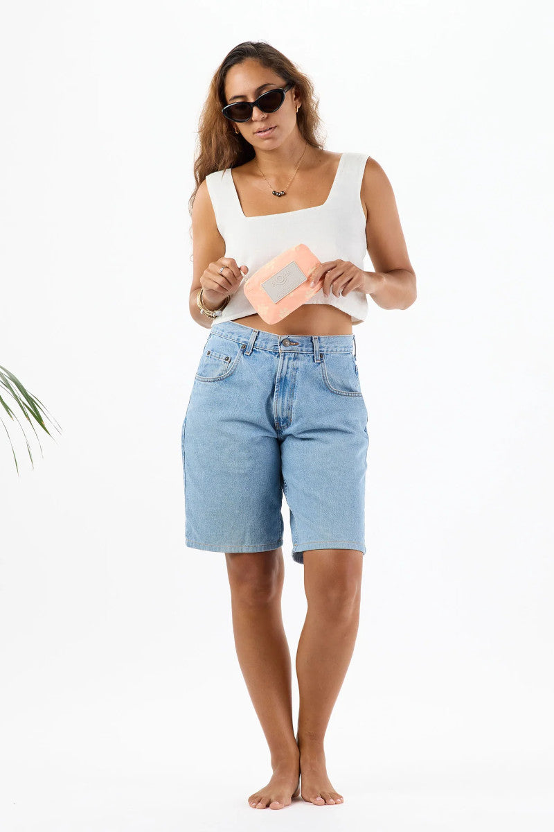 Woman wearing a white crop top and blue shorts holding a pink floral pouch on a white background