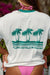 Back of a woman wearing a Billabong t-shirt with a palm tree design and pink bikini bottoms outdoors.