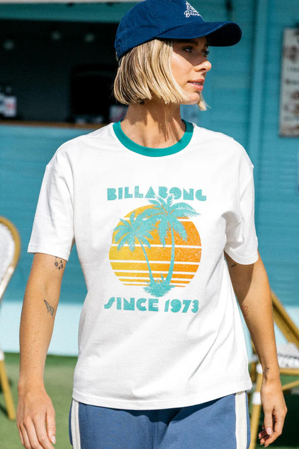 Woman wearing a white t-shirt with a Billabong logo and design, standing against a blue wall.