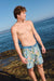 Man wearing colorful swim shorts standing on a rocky beach with ocean in the background