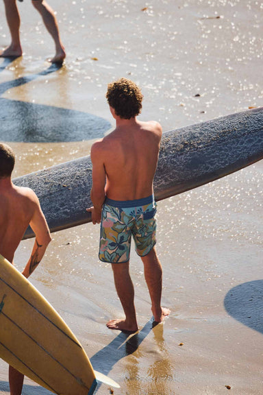 Two people with surfboards on a beach, one wearing colorful swim shorts.