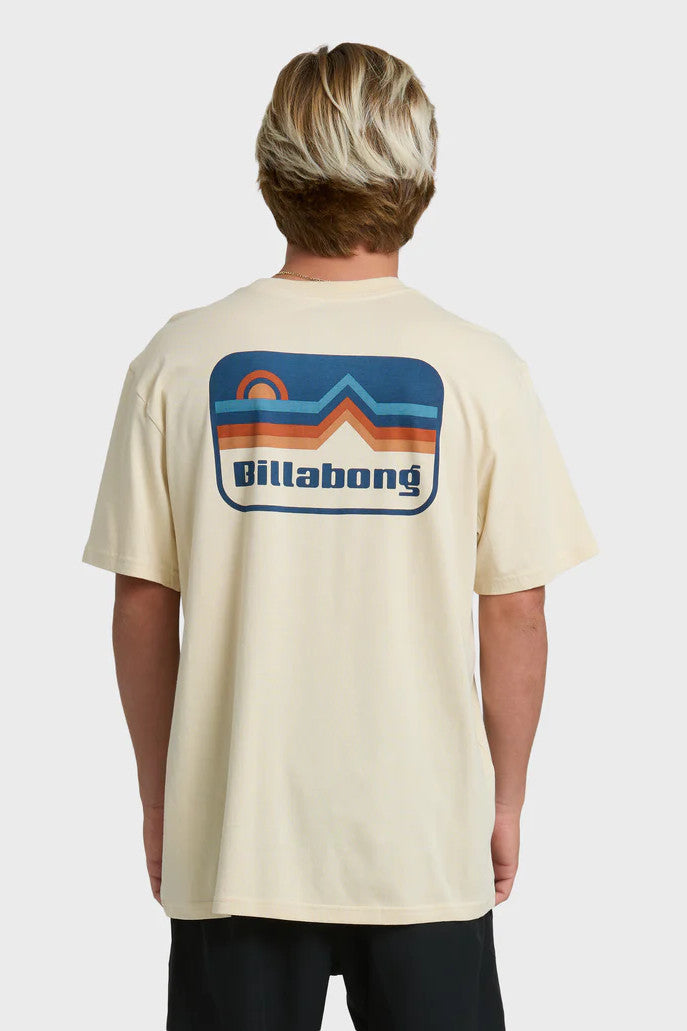 A man wearing a beige t-shirt with short sleeves, featuring a blue and orange mountain and sun graphic on the back.