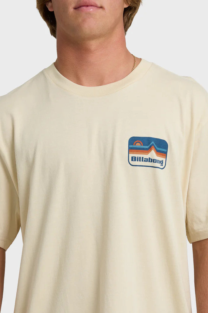 A man wearing a beige t-shirt with short sleeves, featuring a blue and orange mountain and sun graphic on the chest.