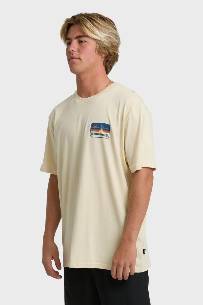 A man wearing a beige t-shirt with short sleeves, featuring a blue and orange mountain and sun graphic on the chest.