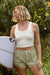 Woman holding a surfboard in white tank top and green drawstring shorts in front of trees