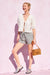Woman in a white lace top and striped shorts holding a brown bag against a pink background