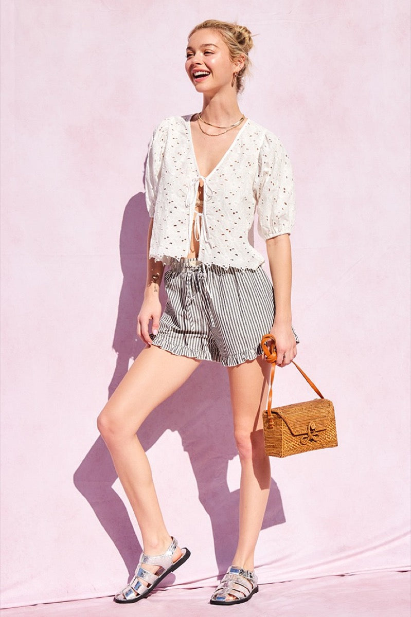 Woman in a white lace top and striped shorts holding a brown bag against a pink background