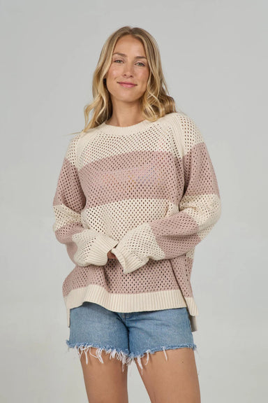 Woman wearing a loose knit sweater with cream and taupe colorblock stripes over denim shorts.