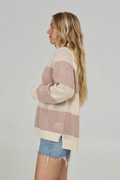 Side view of a woman wearing a loose knit sweater with cream and taupe colorblock stripes over denim shorts.