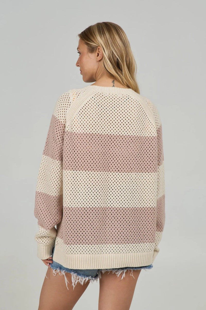 Back view of a woman wearing a loose knit sweater with cream and taupe colorblock stripes over denim shorts.