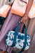Person holding a dark teal floral-patterned duffel bag with another bag in the background.
