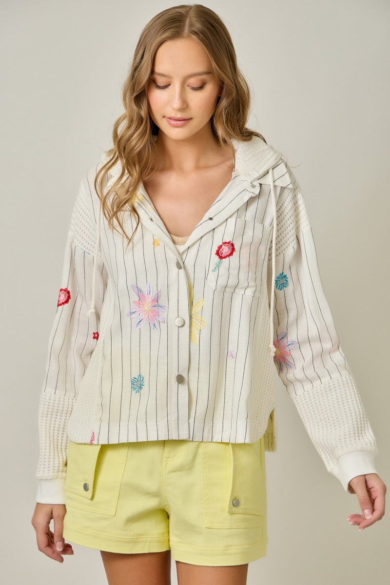 Woman wearing a light-colored embroidered jacket with colorful flowers and yellow shorts on a neutral background