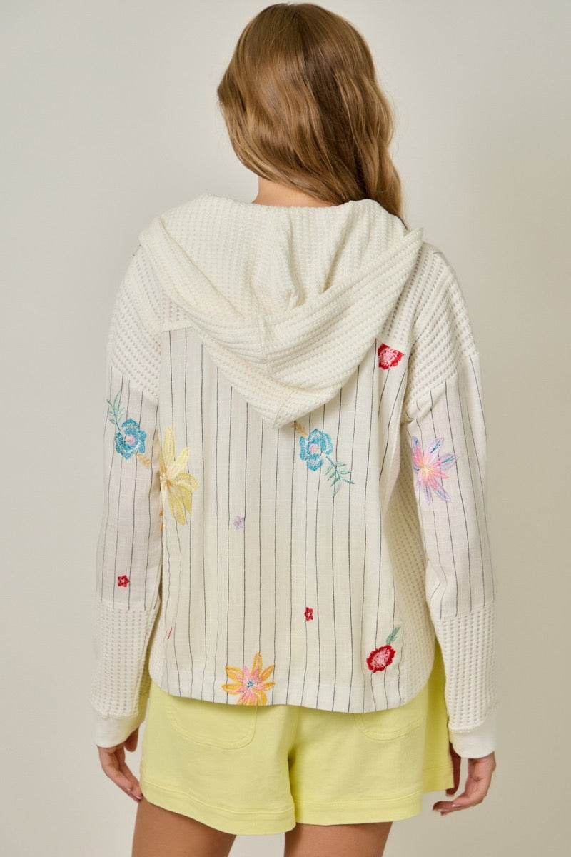 Woman wearing a white jacket with colorful floral embroidery and yellow shorts on a plain background