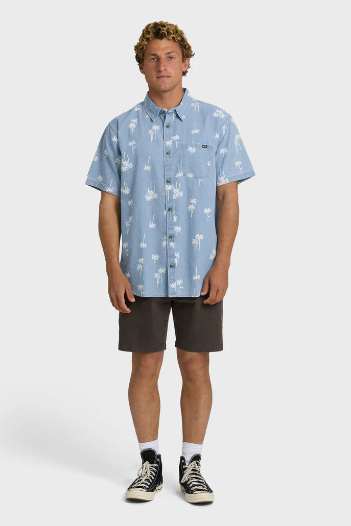 Man wearing a light blue button-up shirt with white palm tree pattern and dark shorts on a white background
