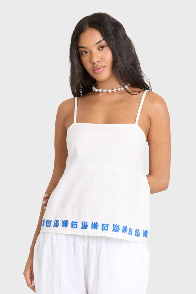 Woman wearing a white tank top with blue embroidery border on a plain background