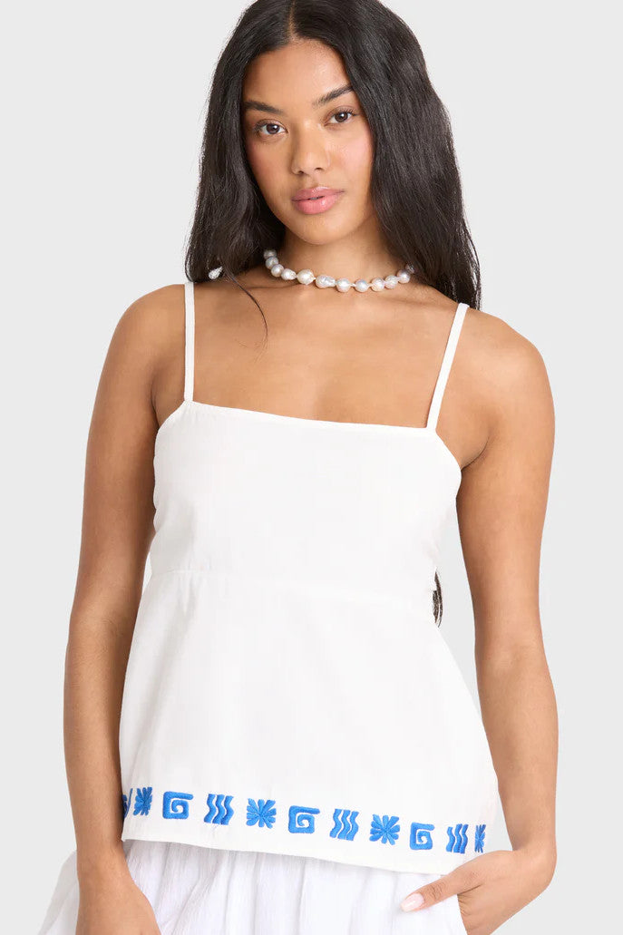 Woman wearing a white tank top with blue embroidery border on a plain background