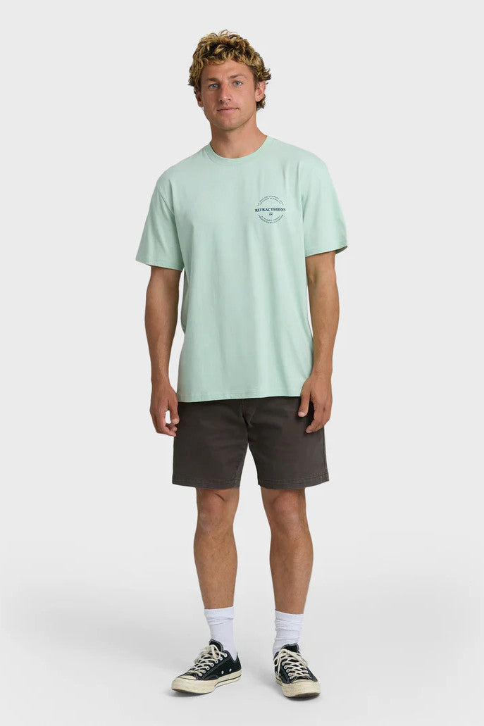 Man wearing a light green t-shirt with a chest logo and dark shorts on a white background