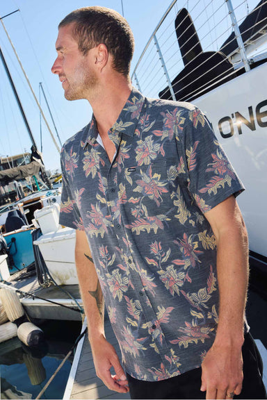 Man wearing a floral patterned shirt on a dock with boats in the background