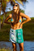 Woman in a blue bikini top and green skirt with palm tree design standing by a body of water.