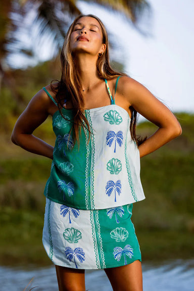 Woman wearing a green and white tank and skirt with palm tree patterns by a body of water.