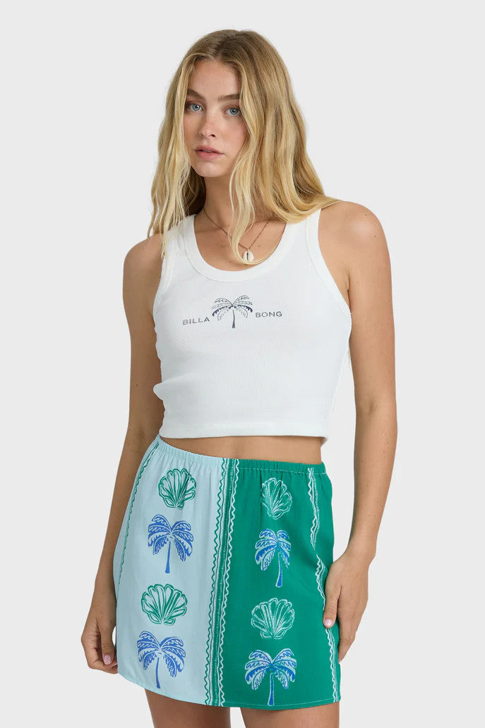 Woman wearing a white tank top with palm tree design and a green and blue skirt with leaf patterns on a white background.