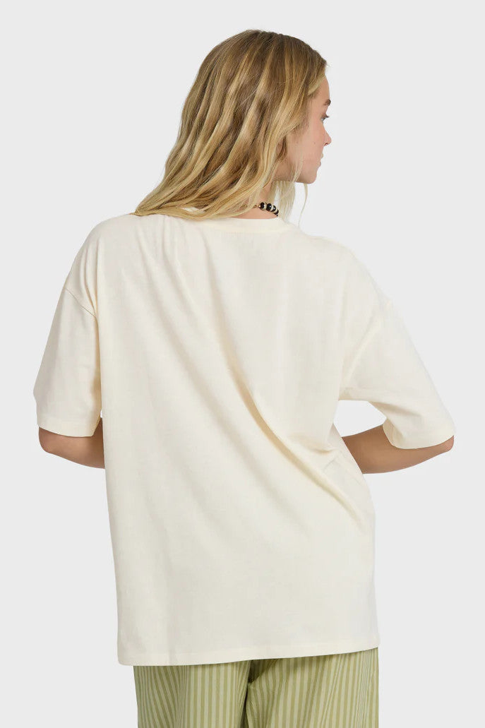 Back view of a woman wearing a cream t-shirt with a palm tree and surf shack graphic design and green striped pants on a white background