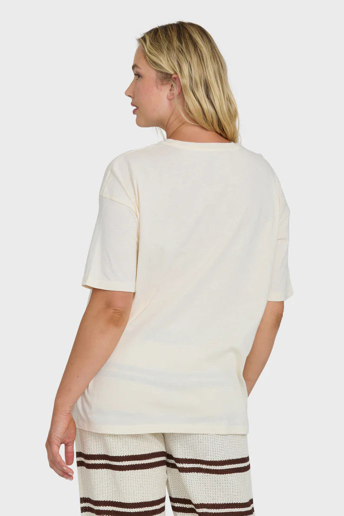 Back view of a woman wearing a cream t-shirt with a palm tree and surf shack graphic design and striped pants on a white background